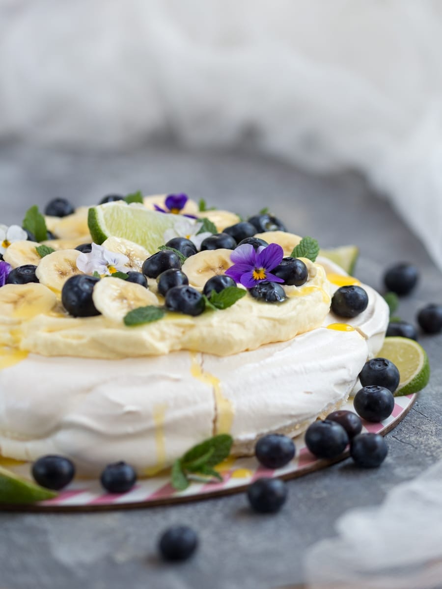 Australian cake pavlova with mango cream, blueberries, banana slices, pansies, mint leaves and lime curd drizzle.