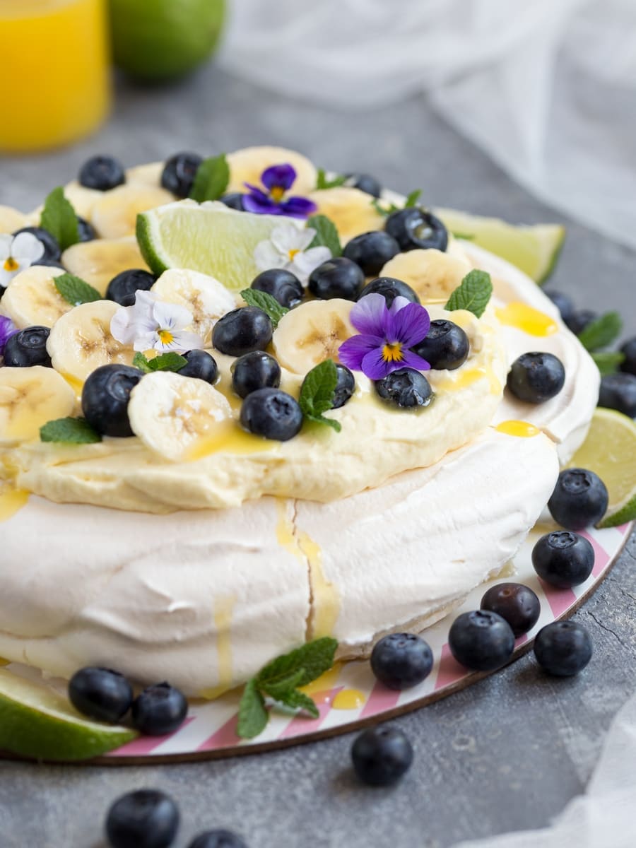 Australian cake pavlova with mango cream, blueberries, banana slices, pansies, mint leaves and lime curd drizzle.