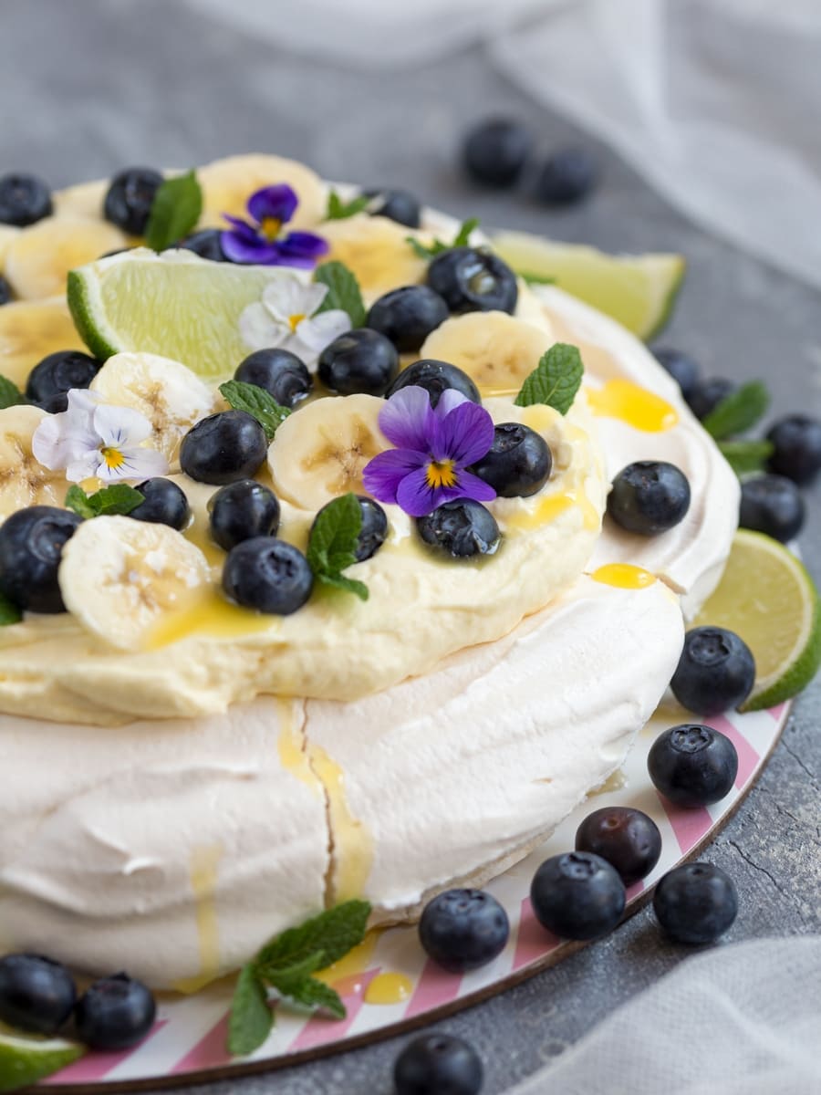 Australian cake pavlova with mango cream, blueberries, banana slices, pansies, mint leaves and lime curd drizzle.