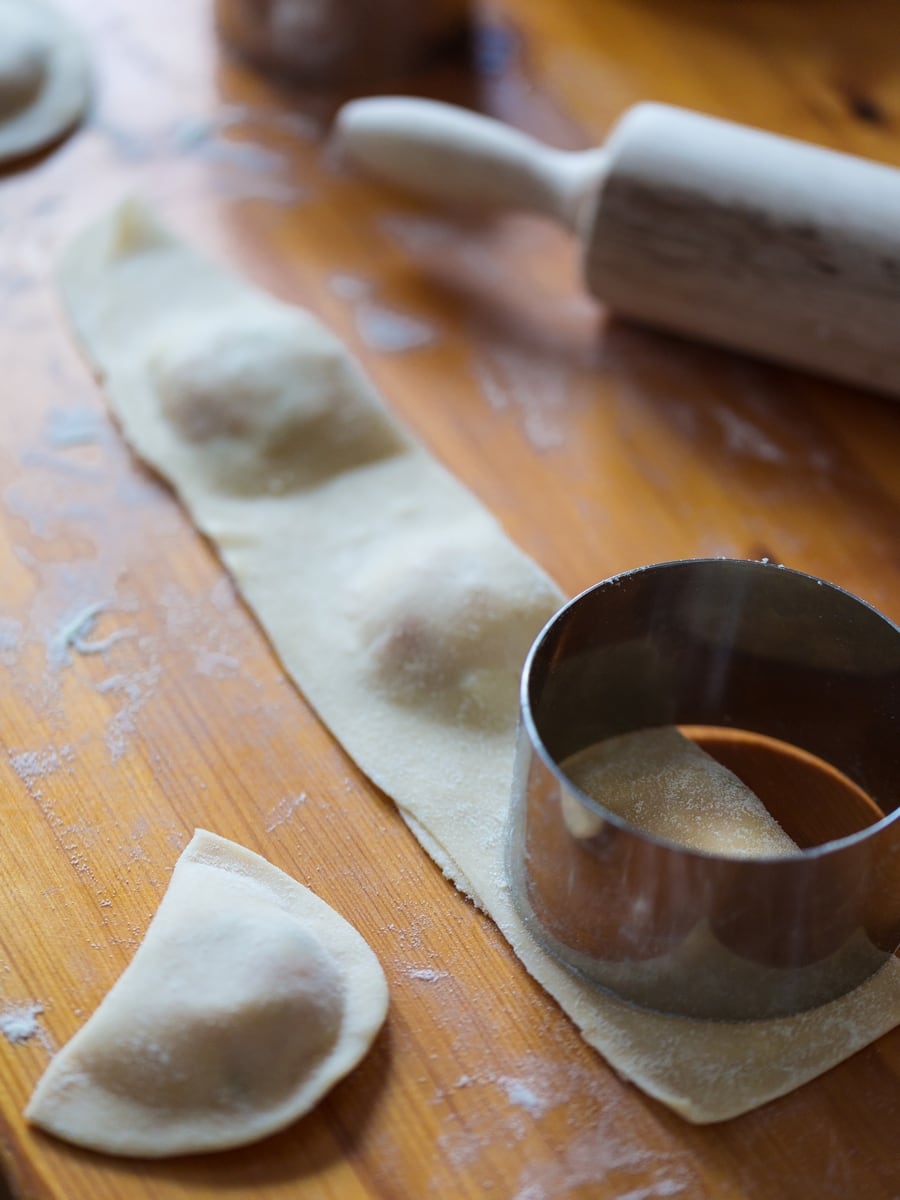 Pastry ring cutting out mezzelune ravioli from filled pasta dough on wooden surface.
