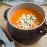 Roasted pepper soup topped with crumbed goat cheese in a brown clay pot.