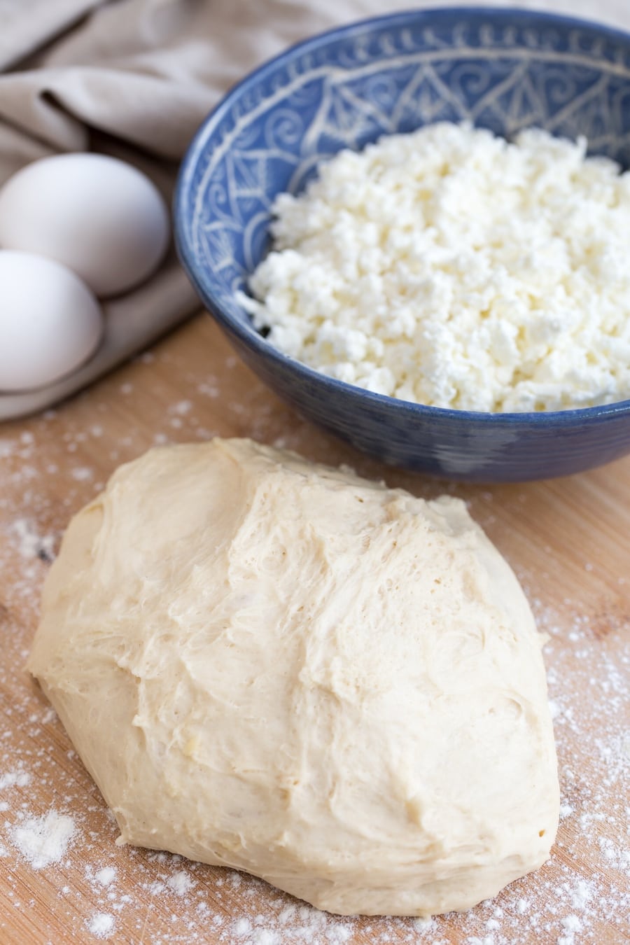 Ball of dough on floured wooden board, bowl with shredded cheeseand eggs in the background.