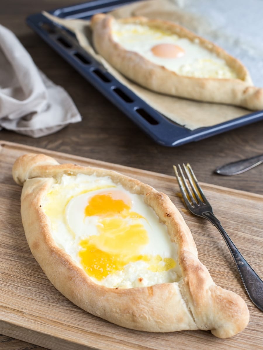 Georgian bread khachapuri adjaruli, one on wooden board, one on oven tray.