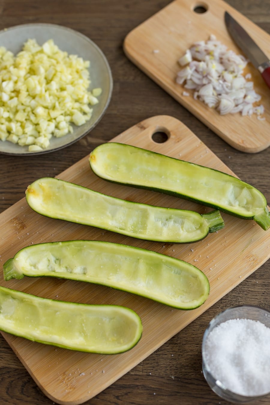 Carved out zucchini halves on wooden board, other chopped ingredients laid around.