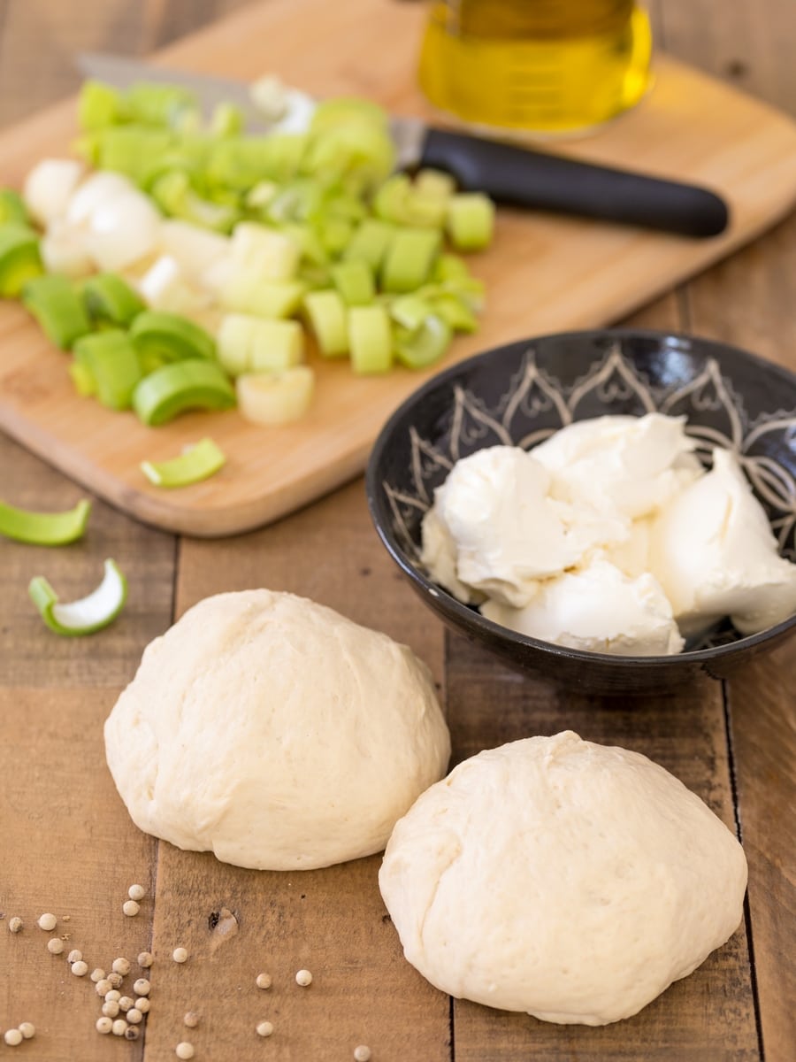 Two balls of pizza dough, mascarpone in a bowl, chopped leek on a cutting board.
