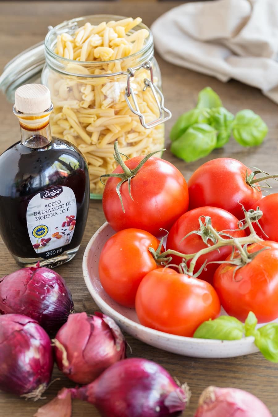 Ripe tomatos in a bowl, red onions around it, a bottle of balsamico vinegar and strozzapreti pasta in a jar.