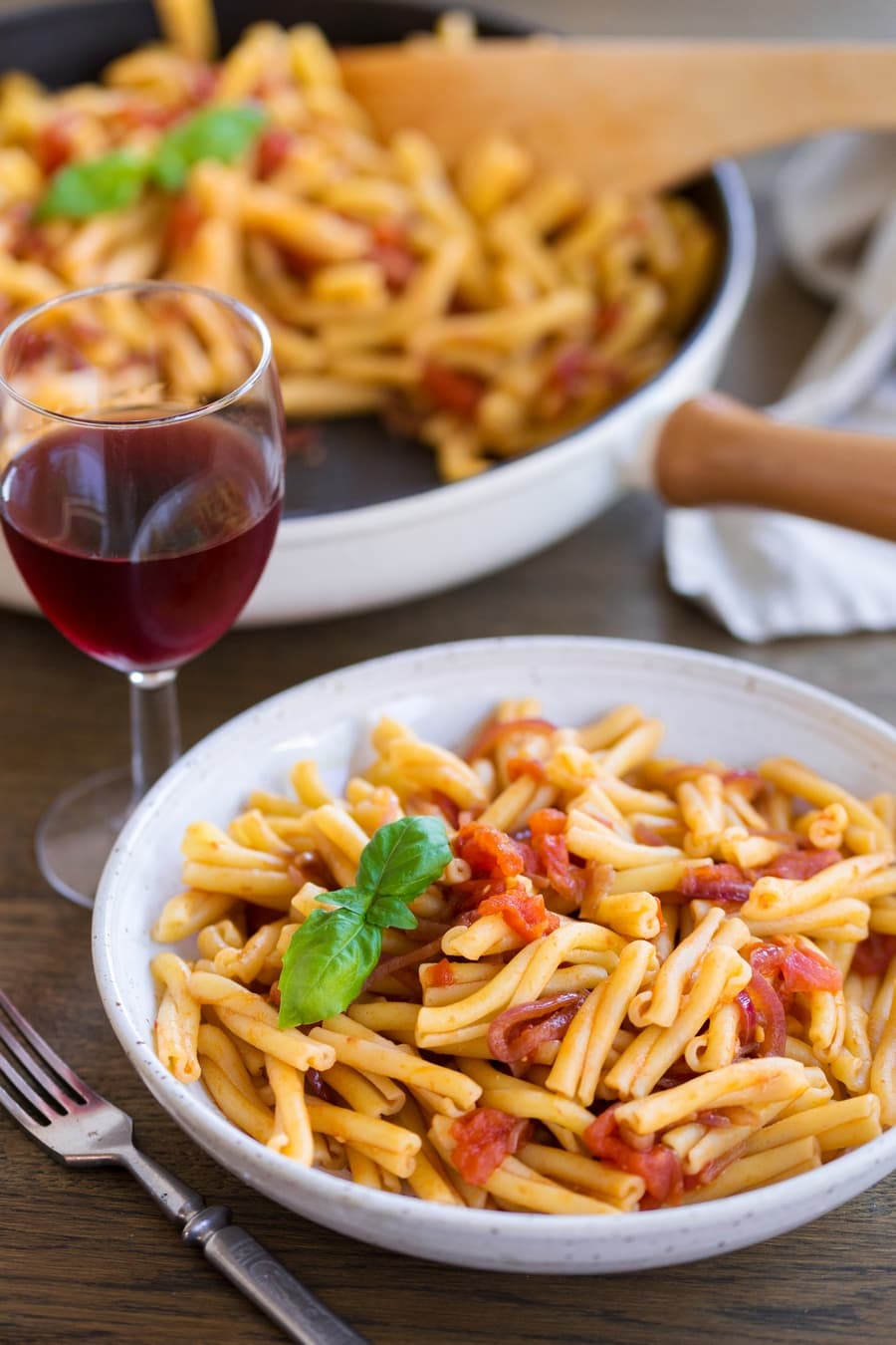 Balsamico pasta with onions and tomatoes served in a bowl, pan containing same food in the background, glass of red wine.