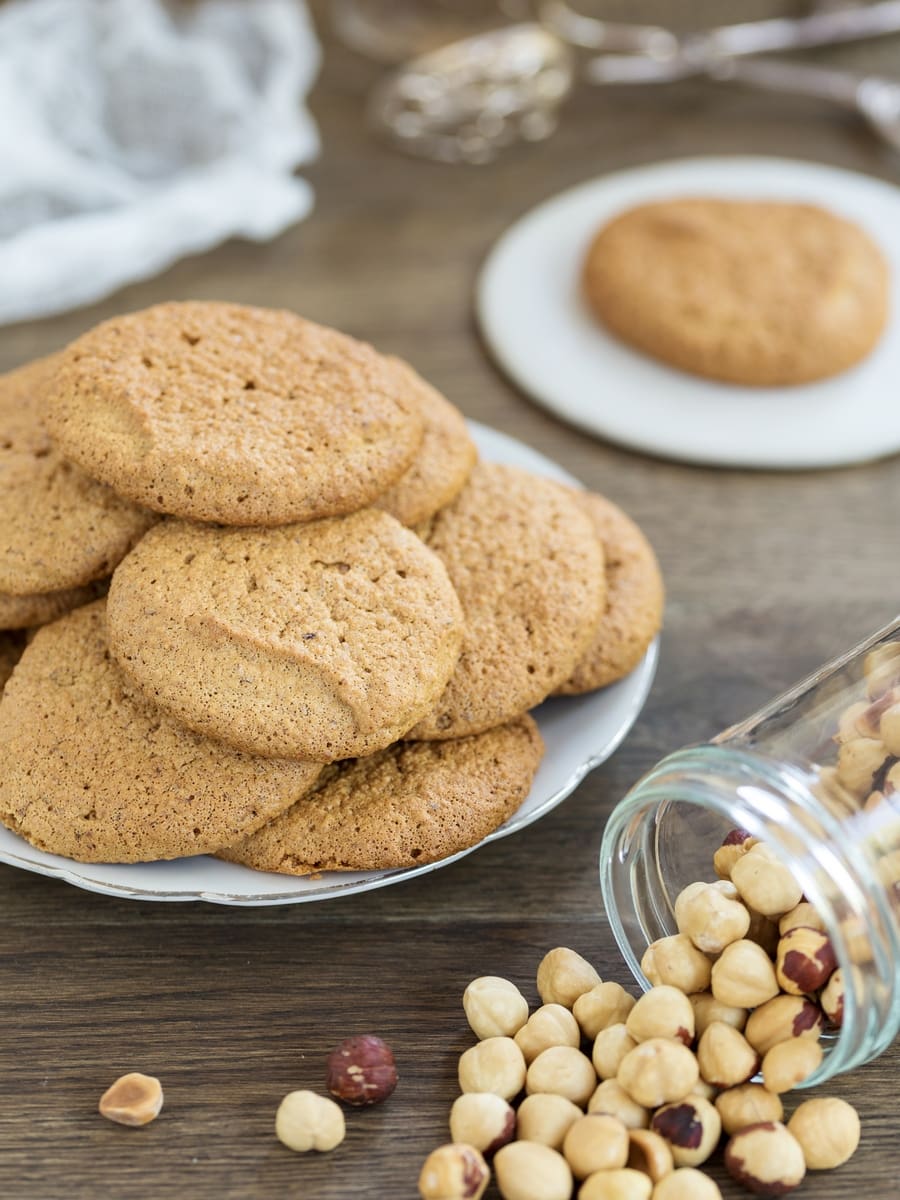 Hazelnuts pouring out of a tilted jar, hazelnut honey meringue cookies stacked on a plate.