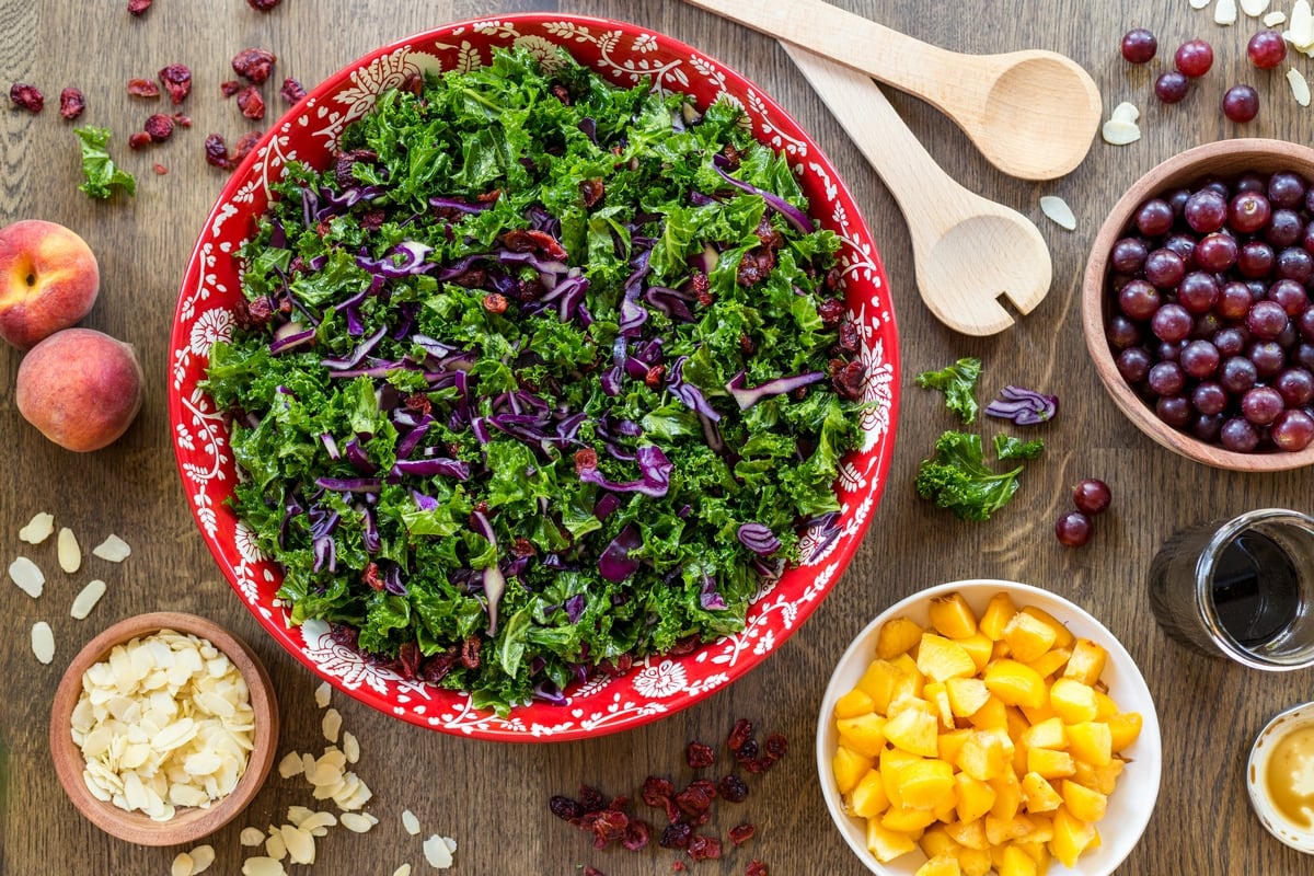 Flatlay view of kale salad in a red bowl. Small bowl with chopped fruits placed around it.
