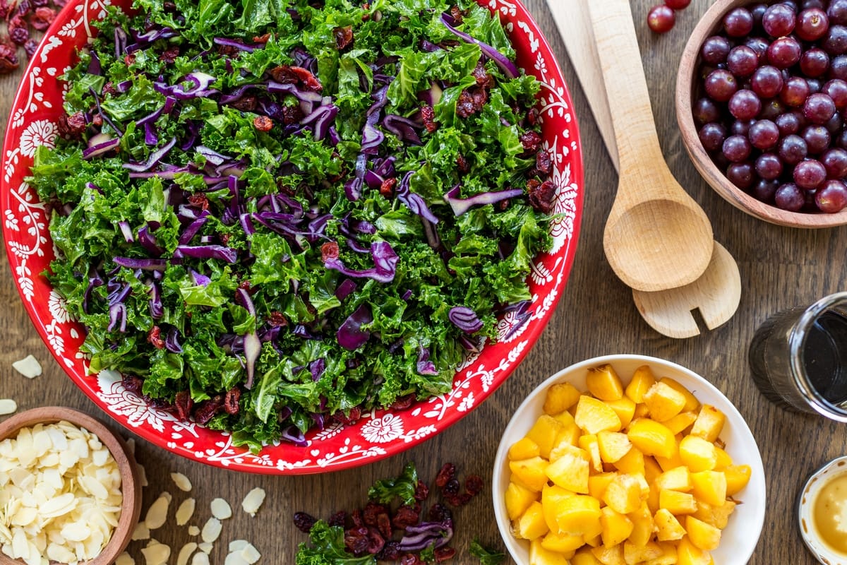 Flatlay view of fruity kale salad in a red bowl. Small bowl with chopped fruits placed around it.