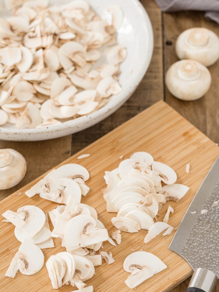Cultivated mushrooms thinly sliced on a bamboo chopping board, sliced mushrooms in a bowl.