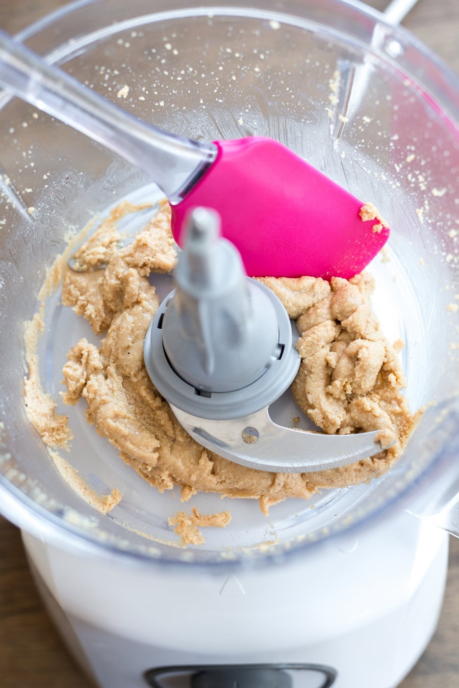 Pink spatula scraping hazelnut paste down the sides of a food processor.