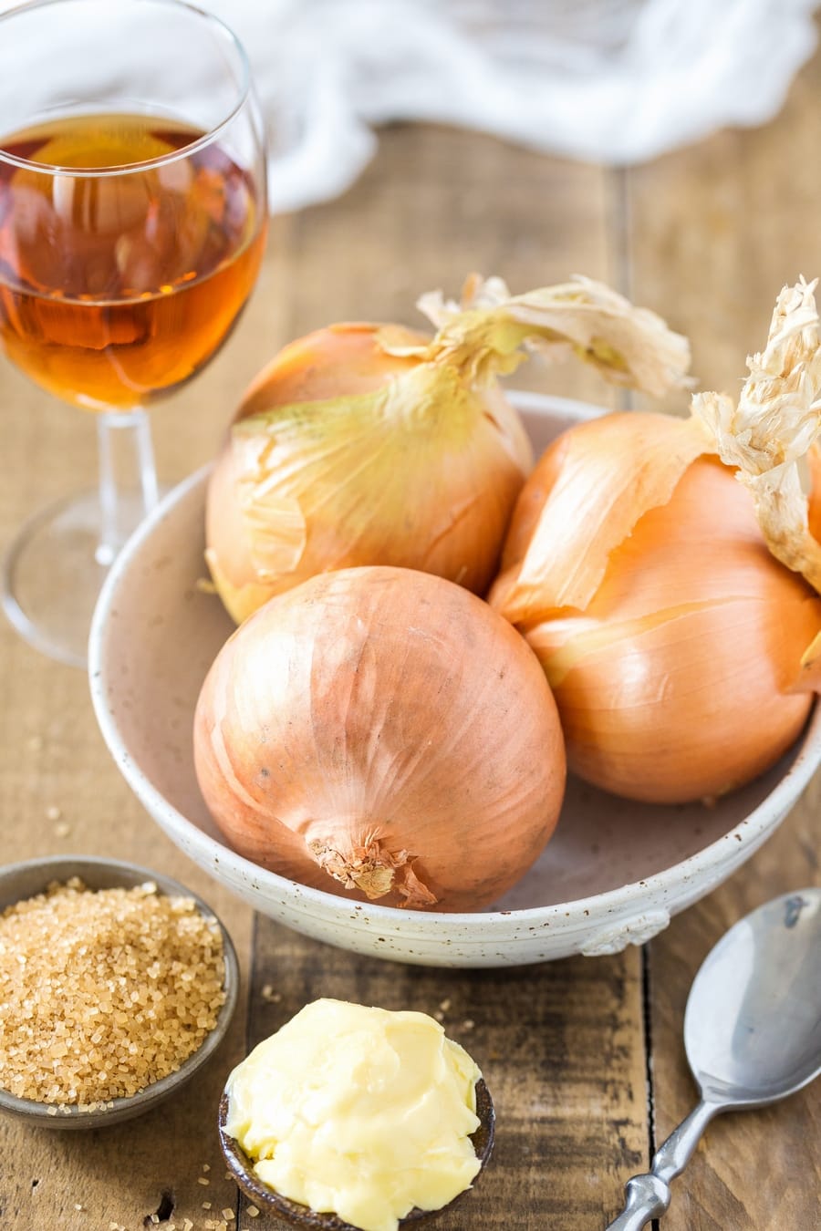 Three yellow onions in a bowl, small bowl of butte rand of brown sugar, glass of marsala in the backgrund. The ingredients to make marsala caramelized onion.