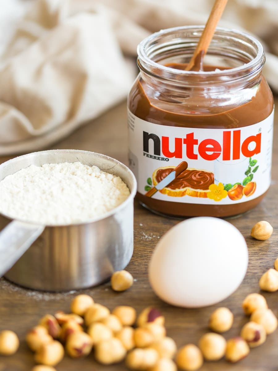 Nutellotti ingredients on a table: one egg, flour, Nutella and whole hazelnuts.