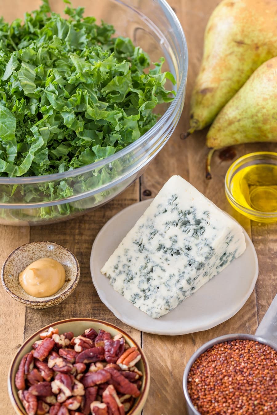A block of blue cheese surronded by kale, pecans, pears, quinoa and condiments in bowls.