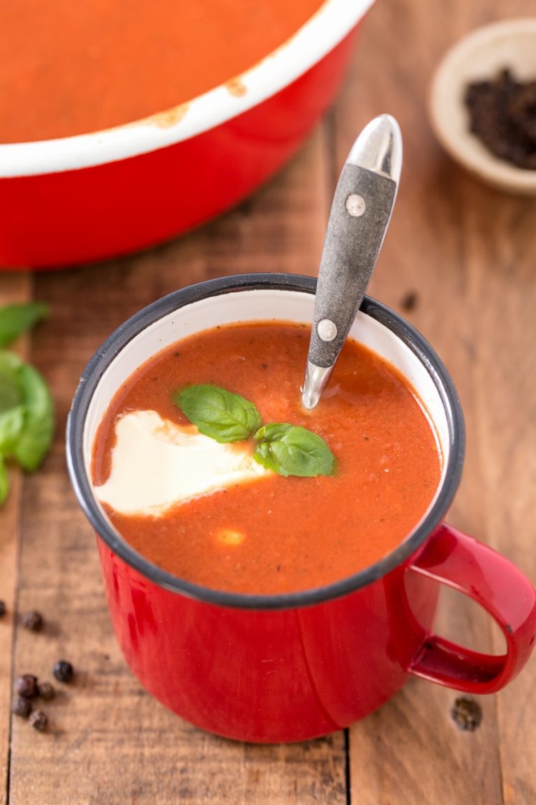 Red metal cup containing pomidorowa soup with a dollop of cream and basil leaves.