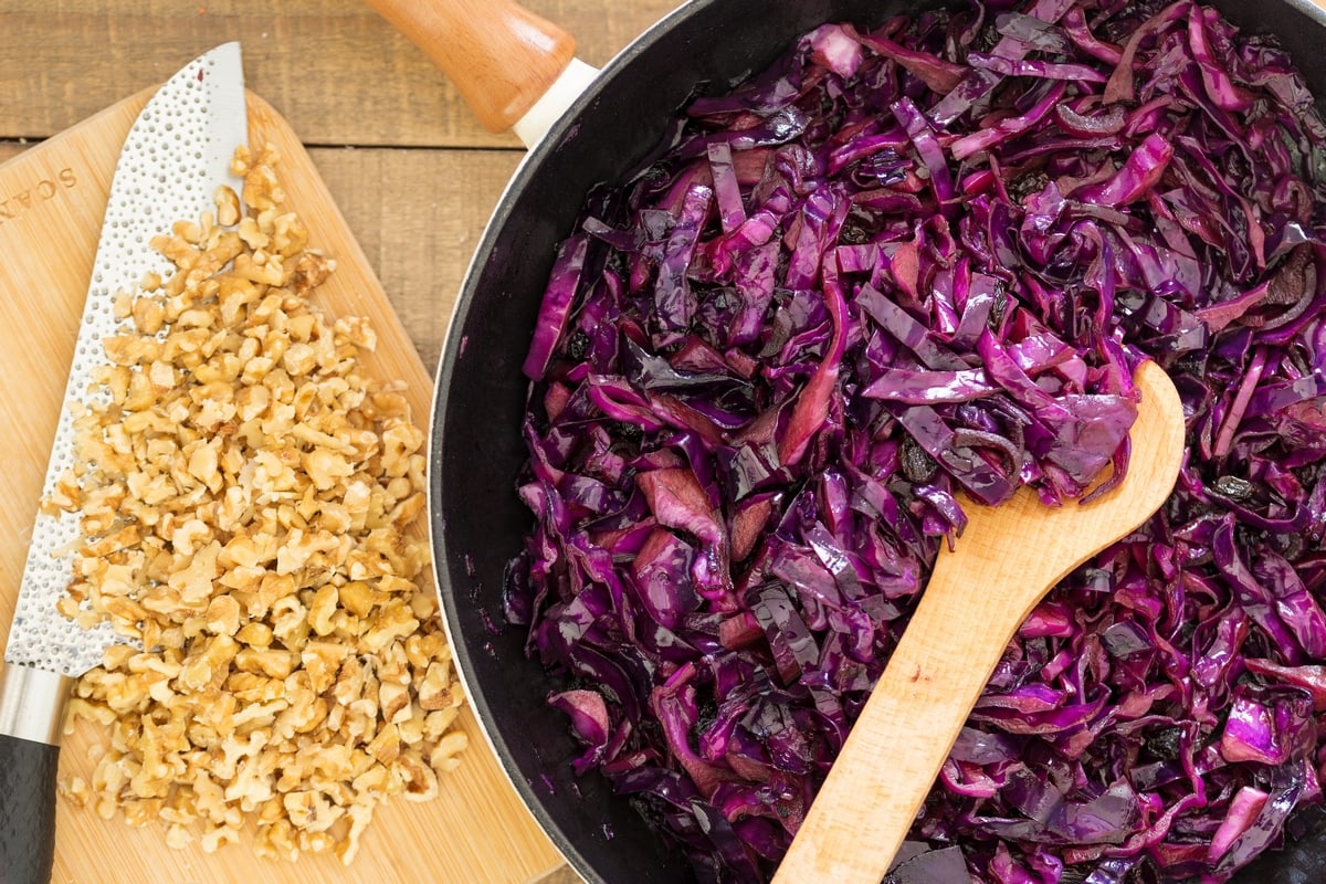 Sautéed red cabbage with onion and raisins in a skillet, chopped walnuts on a chopping board.