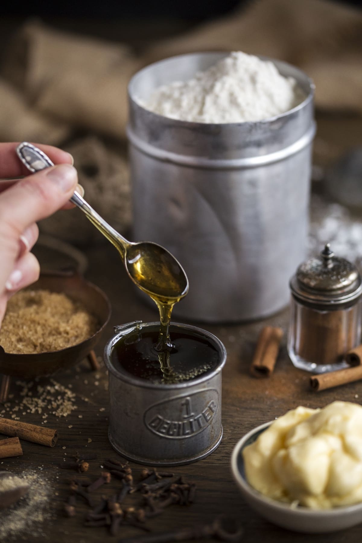 Hand lifting a teaspoon from a container with syrup, other recipe ingredients placed around it.
