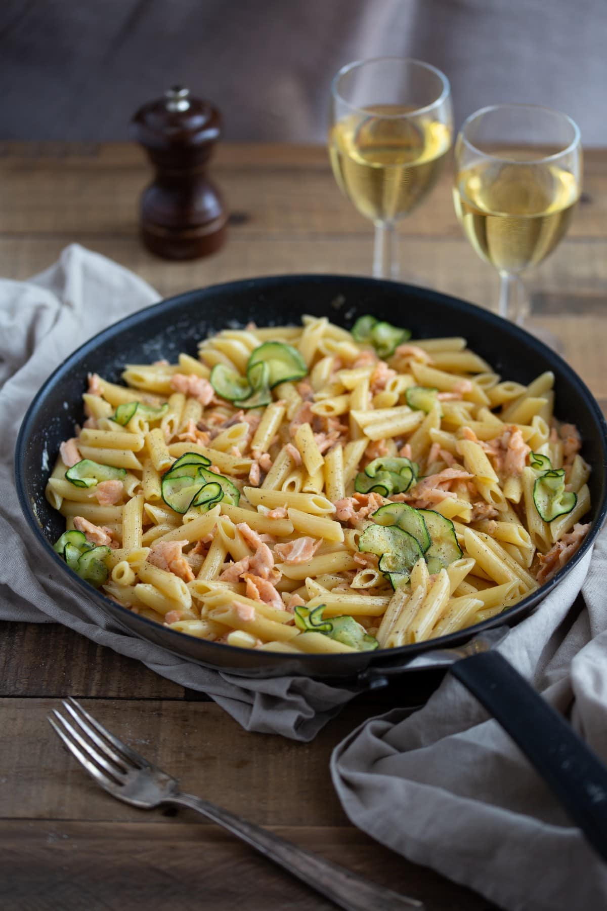 A pan filled with salmon pasta next to two wine glasses.