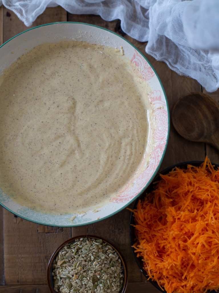 Cake batter in a bowl, shredded carrots and seeds aside.
