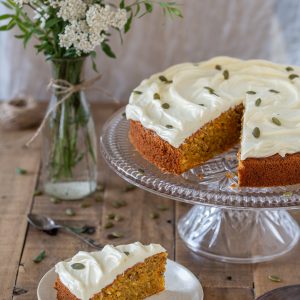 A slice of carrot cake on a plate, the rest of the cake on a cake stand.