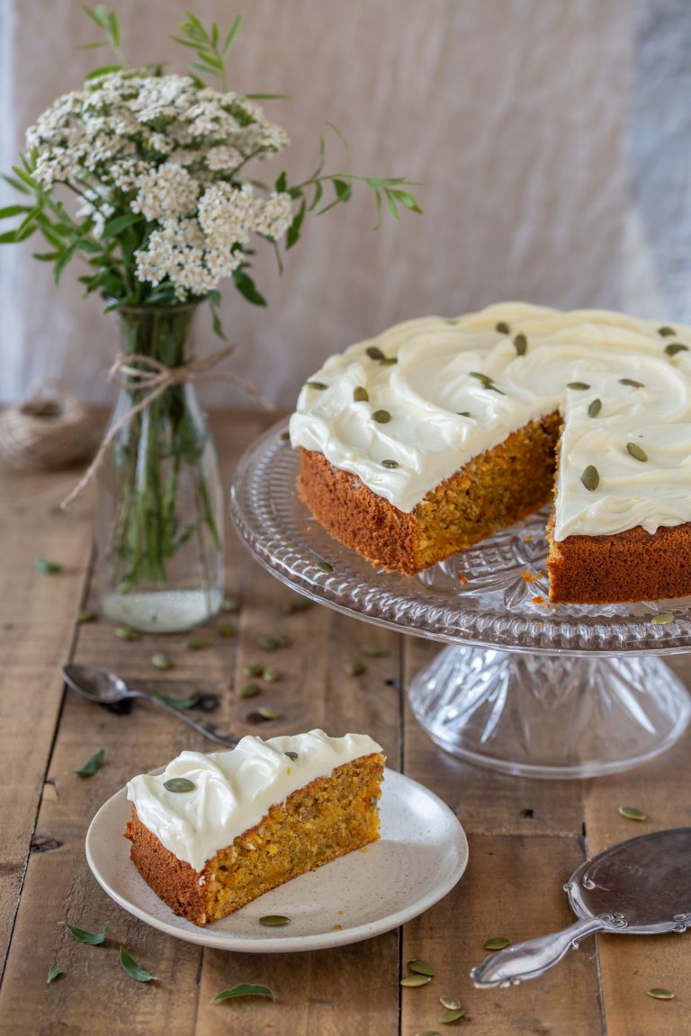 A slice of carrot cake on a plate, the rest of the cake on a cake stand.