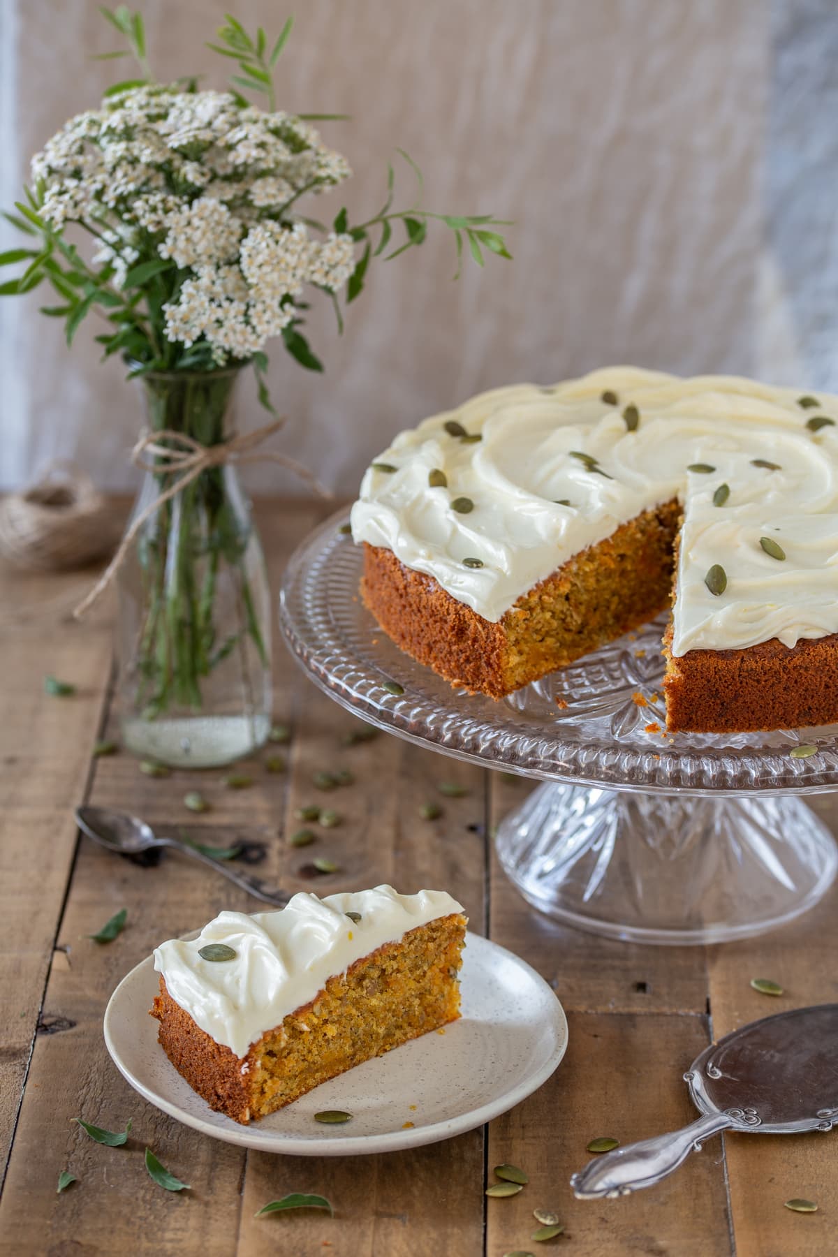 A slice of carrot cake on a plate, the rest of the cake on a cake stand.