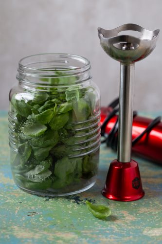 A mason jar filled with basil leaves, immersion blender on the side.