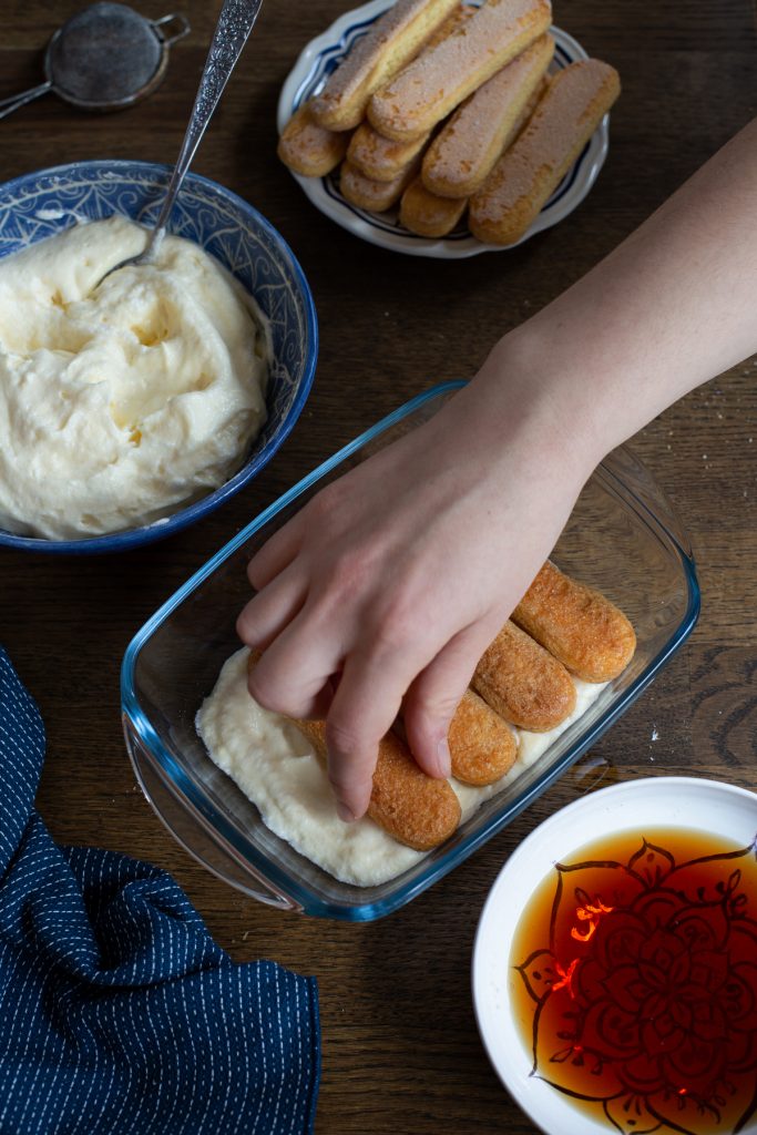 Hand adding soaked ladyfingers over mascarpone cream.
