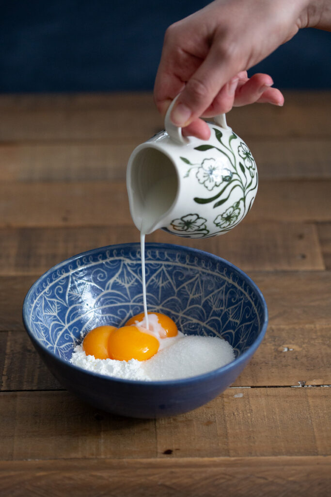 Pouring milk from a jug into a bowl containing sugar, starch and egg yolks.
