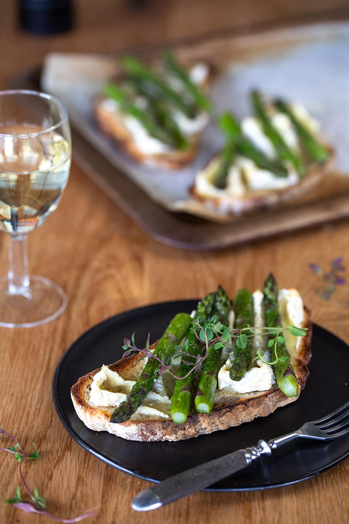 One bruschetta with asparagus and camembert on black plate, more in the background.