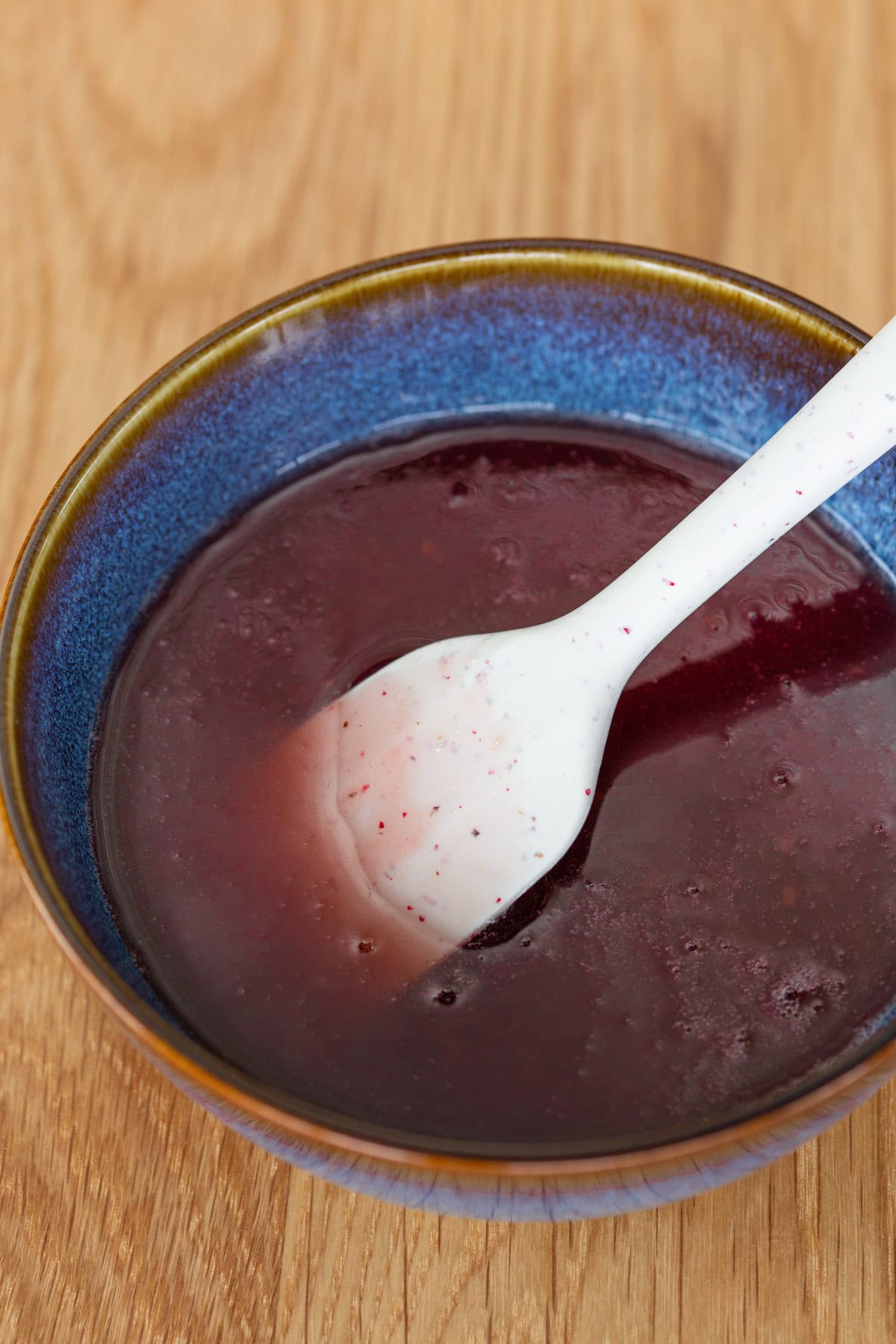 Close-up of raspberry jam in a blue ceramic bowl with a white spatula on a wooden table.
