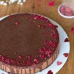 Chocolate raspberry cheesecake covered in chocolate ganache served on a white plate with pink hearts.