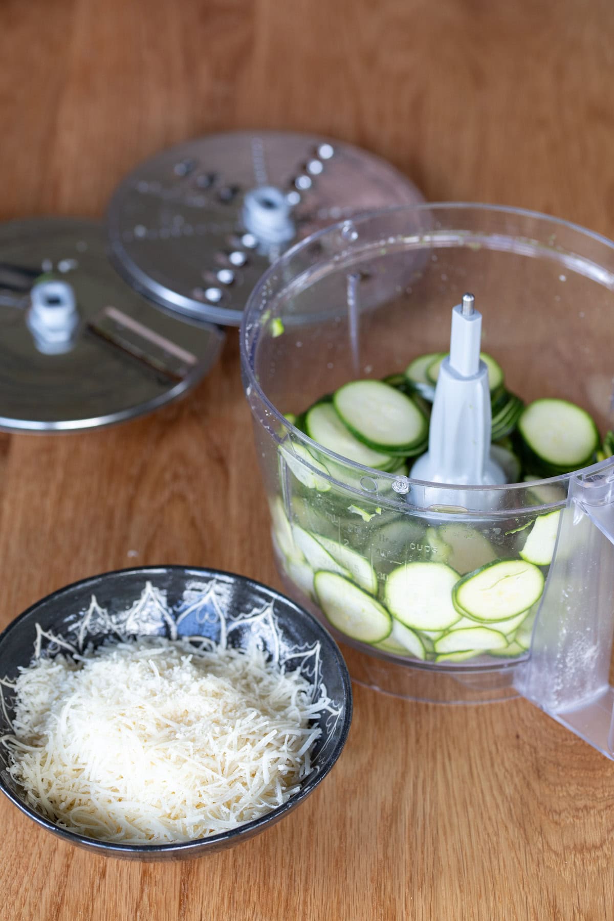 Sliced zucchini in a food processor, grated parmesan in a bowl aside.