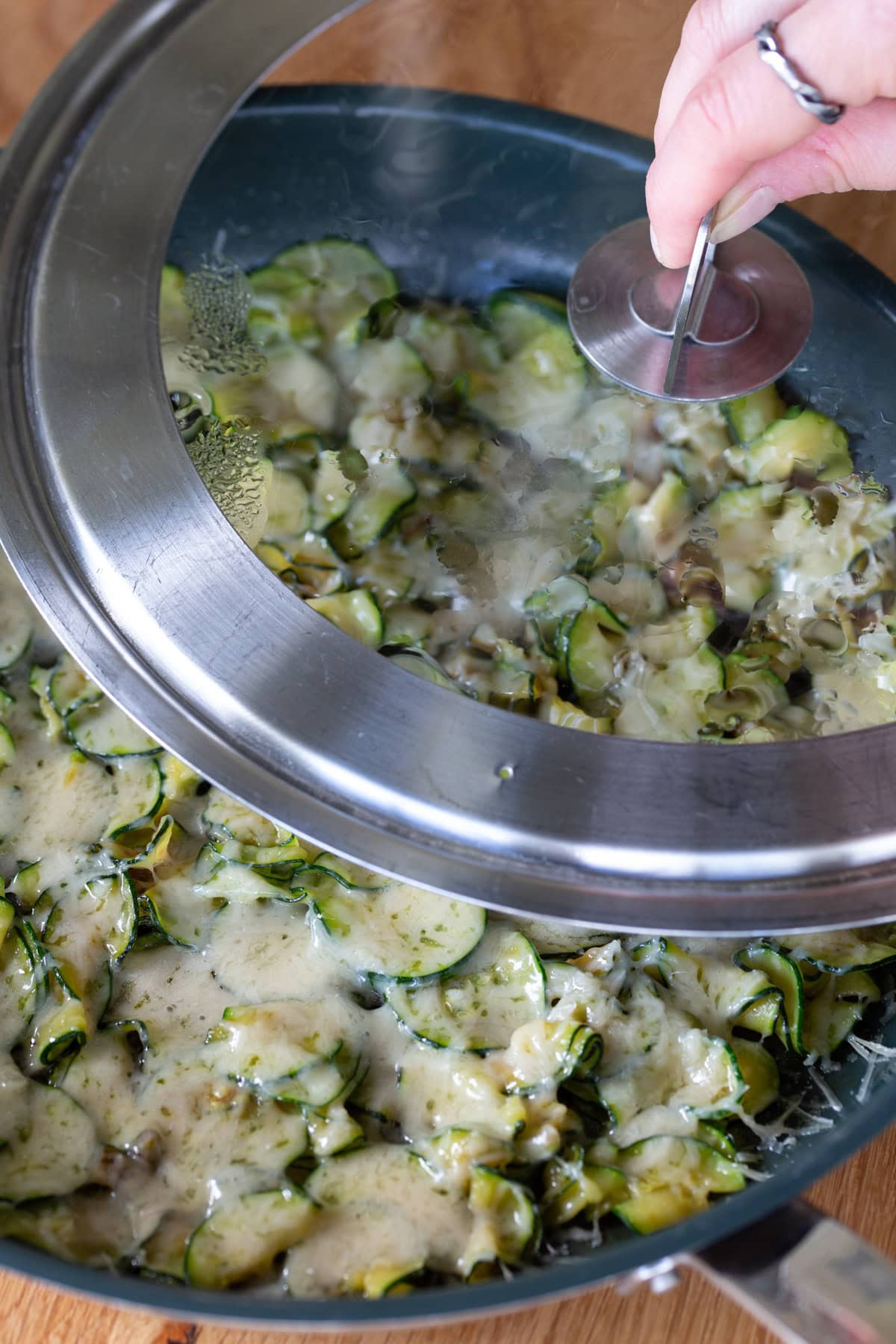 Hand lifting lid to show melted parmesan over sauteed zucchini in a skillet.