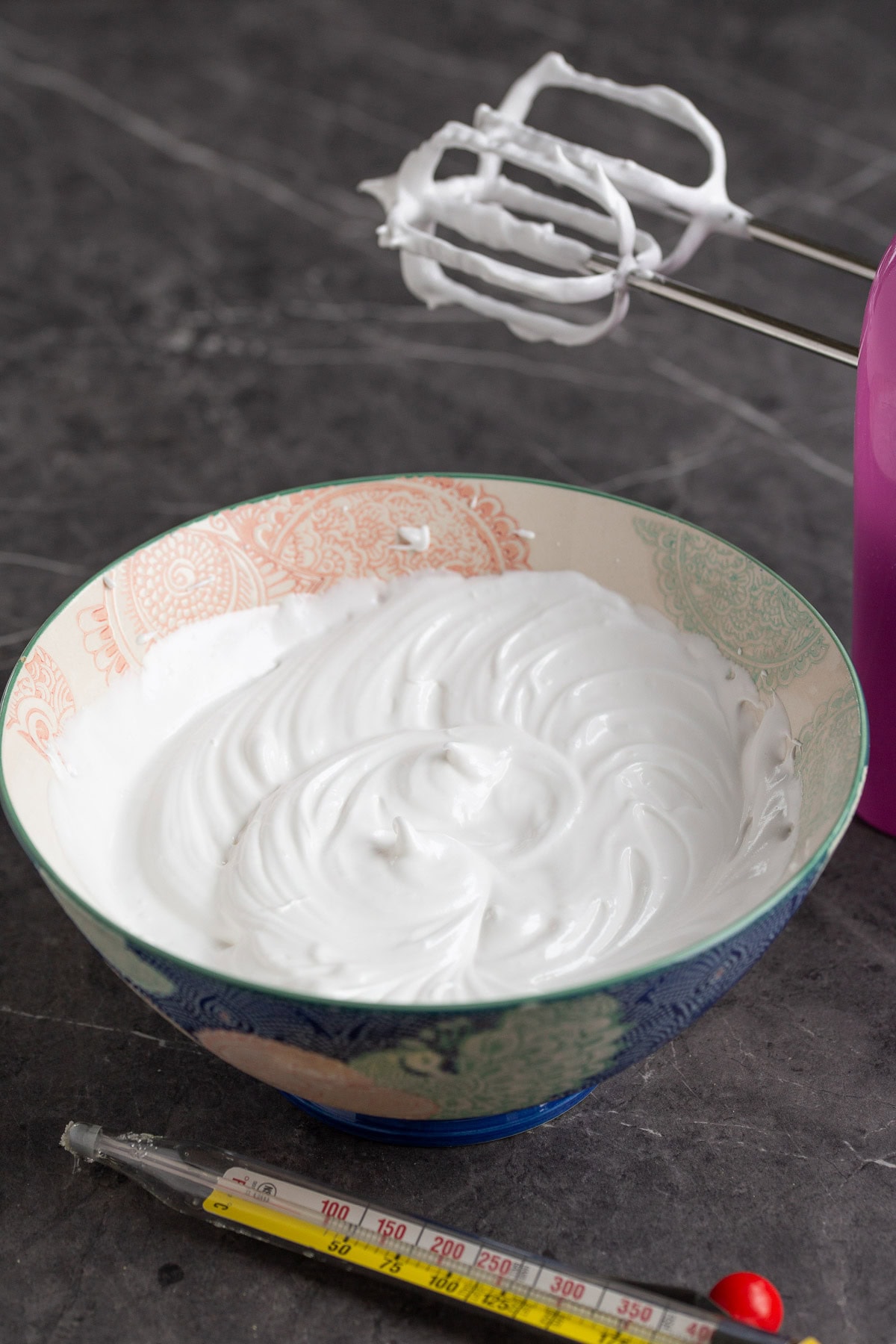 Whipped egg whites in a bowl, candy thermometer in the foreground.