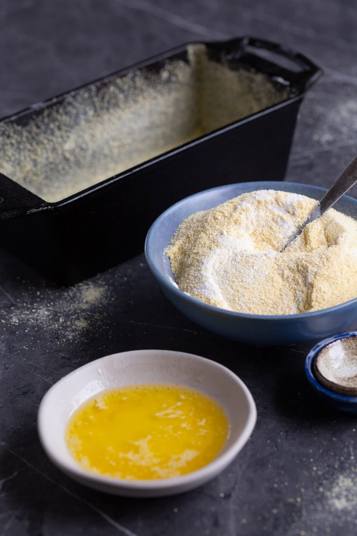 A bowl with melted butter, another bowl with mixed dry ingredients and a greased pan in the background.